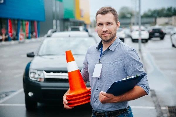 Instruktor autoškoly s tabulkou a dopravním kuželem na parkovišti.
