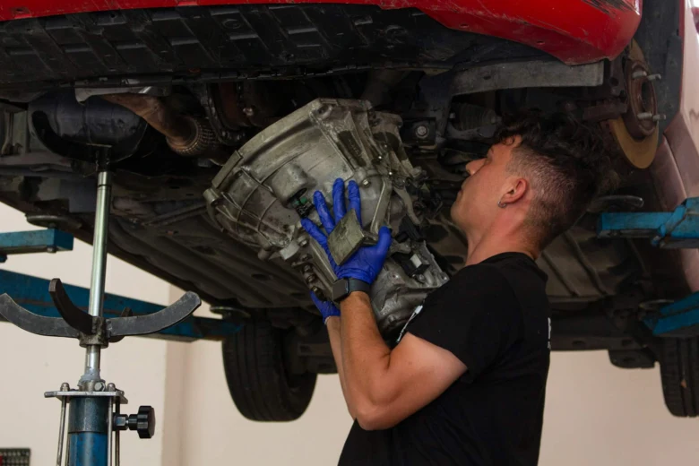 Mechanik drží převodovku pod červeným autem v dílně.