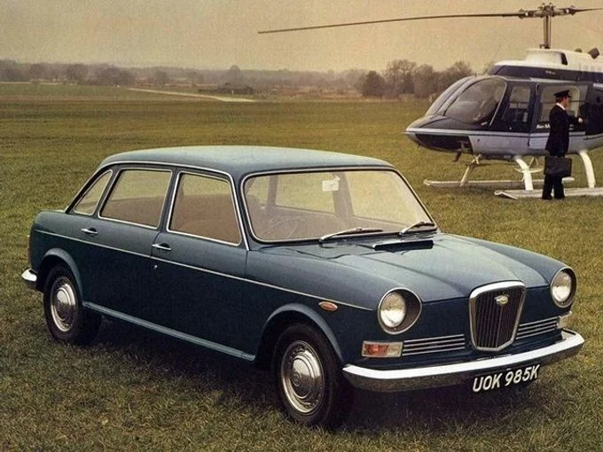 Modrý britský automobil z sedmdesátých let s vrtulníkem na pozadí.