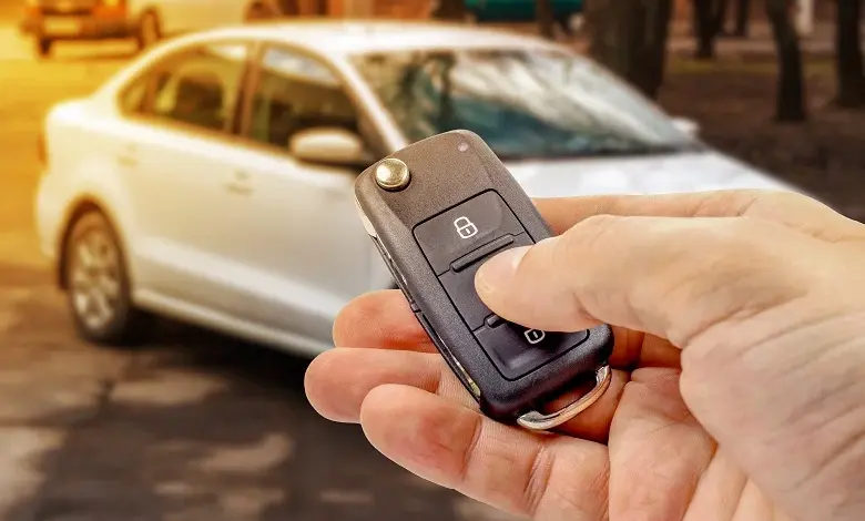 Ruka drží auto klíč s dálkovým ovládáním před bílým autem.