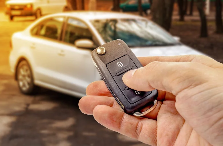 Ruka drží auto klíč s dálkovým ovládáním před bílým autem.