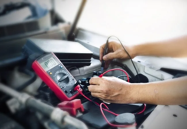 Mechanik měří napětí autobaterie multimetrem pod kapotou auta.