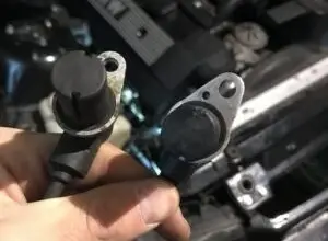 Ruce drží dva snímače vačkového hřídele před otevřeným motorem.