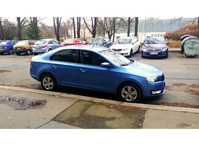 Modrá Škoda Rapid zaparkovaná na parkovišti s jinými auty v pozadí.