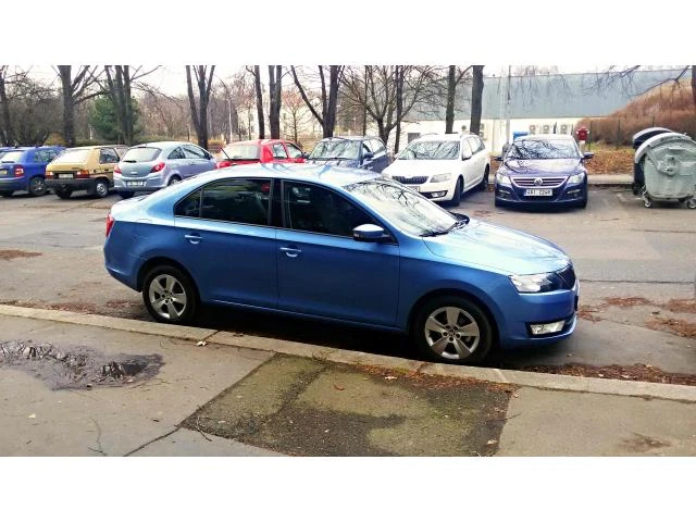 Modrá Škoda Rapid zaparkovaná na parkovišti s jinými auty v pozadí.