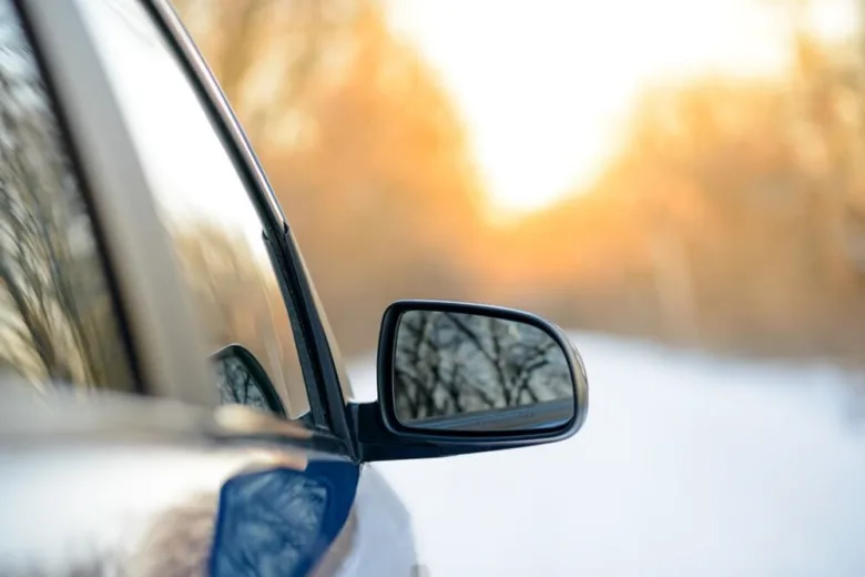 Bílé auto v zimě s odrazem stromů ve zrcátku.