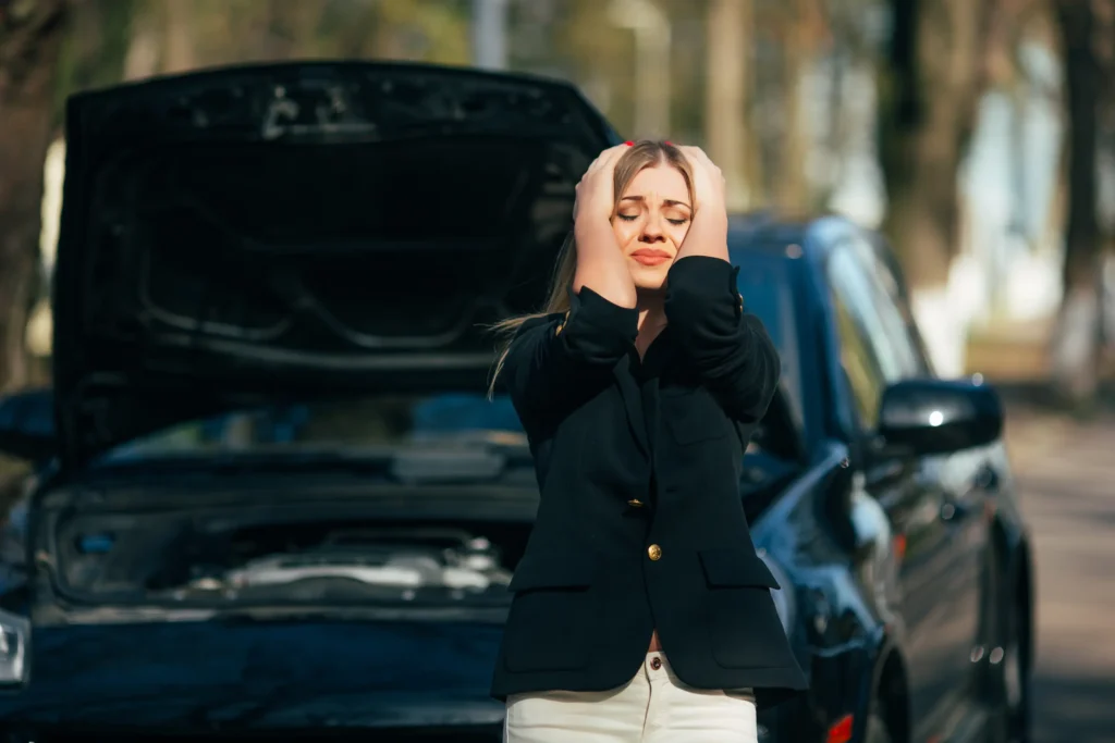 Žena stojí před autem s otevřenou kapotou v parku.
