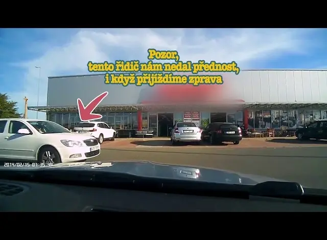 Bílý auto vyjíždí z parkoviště před obchodním domem.