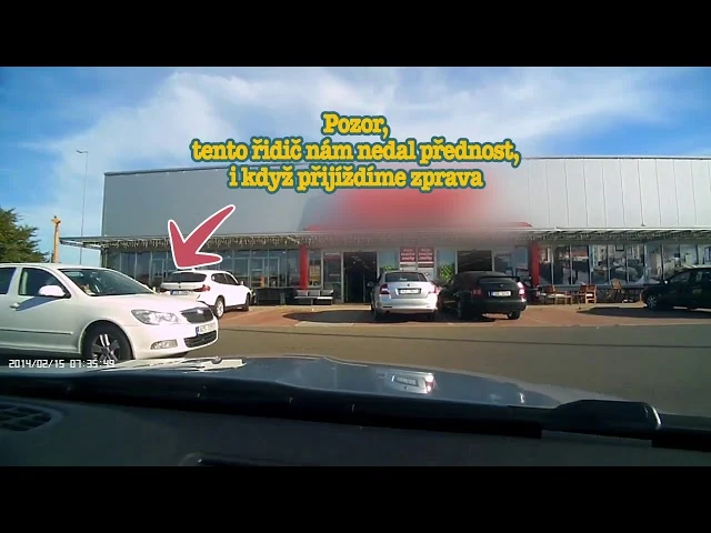 Bílý auto vyjíždí z parkoviště před obchodním domem.