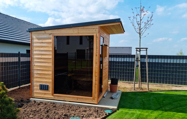 Moderní dřevěný domek s velkými skleněnými panely v zahradním prostředí.