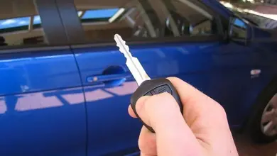 Ruka drží auto klíč před modrým autem na pozadí.