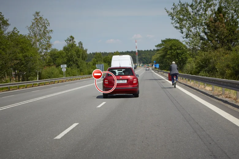 Červené auto jedoucí po silnici s cyklistou na obrubníku.