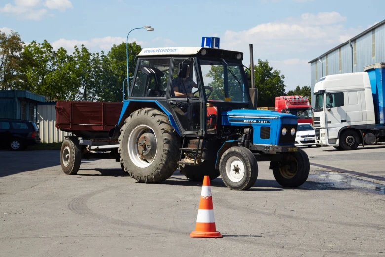 Modrý traktor s návěsem na asfaltové ploše poblíž dopravních značek.