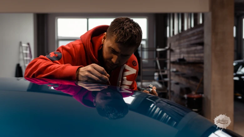 Muž v červené bundě políruje modré auto ve garáži.