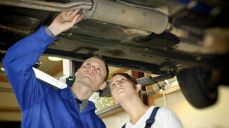 Mechanici kontrolují výfukový systém pod vozem v dílně.
