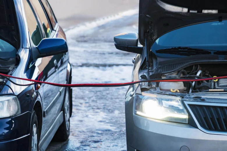 Stříbrné auto s otevřenou motorkou nabíjené červenými kabely od tmavě modrého vozu.