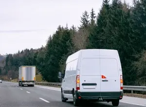 Bílý dodávkový vůz jede za kamionem na dálnici.