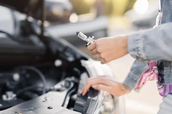 Člověk drží novou žárovku u otevřeného motorového prostoru vozu.