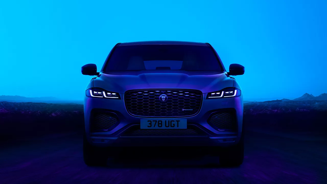 Modrý Jaguar F-Pace SUV s rozsvícenými světlomety před horami.