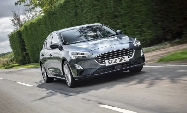 Šedý Ford Focus jezdí po silnici s živým plotem v pozadí.