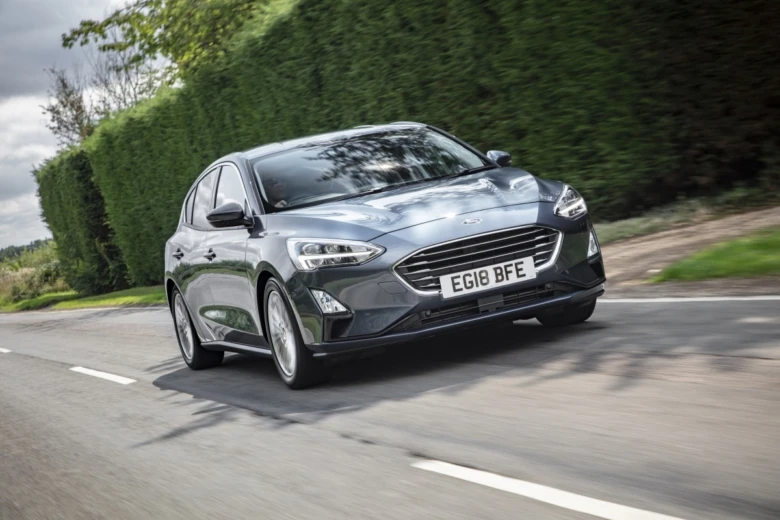Šedý Ford Focus jezdí po silnici s živým plotem v pozadí.