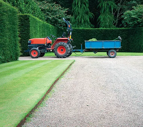 Červený traktor táhne modrý přívěs po zahradní cestě.