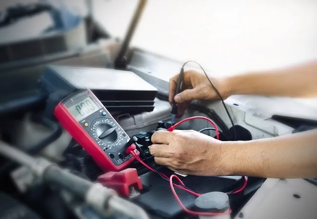 Mechanik měří napětí akumulátoru multimetrem pod kapotou auta.