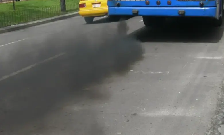 Modrý autobus kouří černým dymem při zrychlení na silnici.