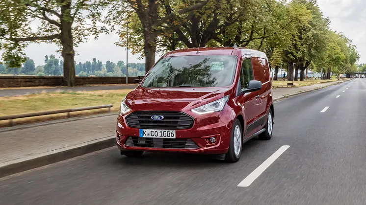 Červený Ford Transit Connect jezdí po silnici pod stromy.