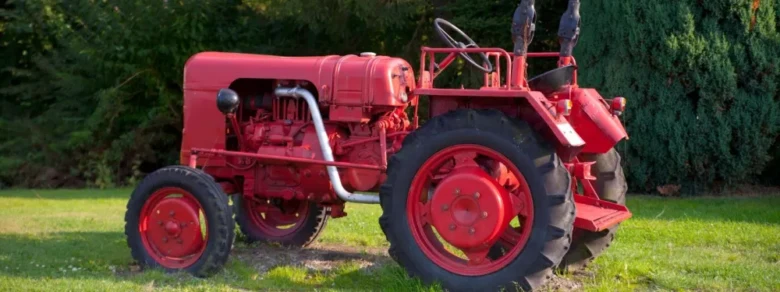 Červený starší traktor stojí na trávníku před stromy.