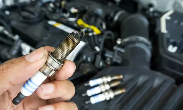 Ruka drží zapalovací svíčku před otevřeným motorem auta.