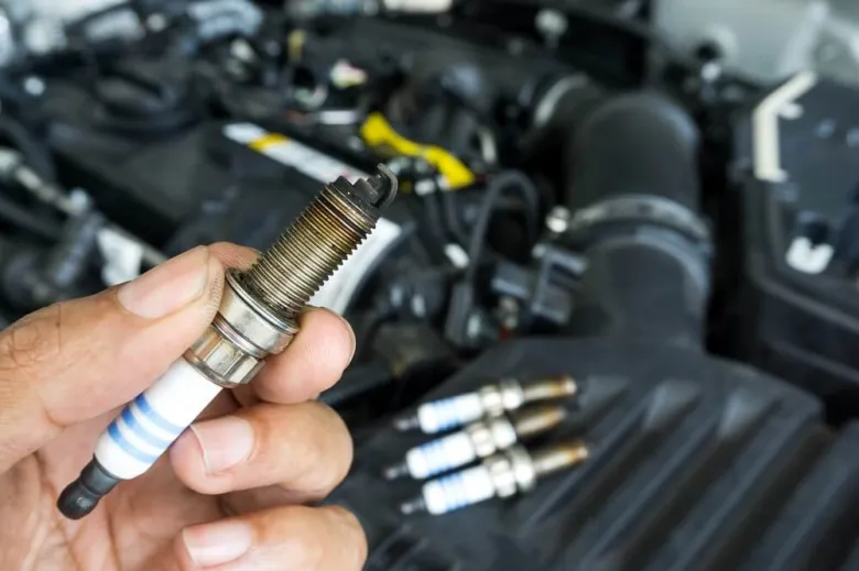 Ruka drží zapalovací svíčku před otevřeným motorem auta.