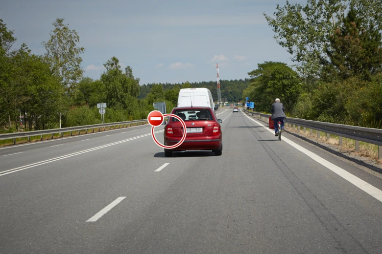 Červený osobní automobil jedoucí po dálnici s cyklistou vpravo.
