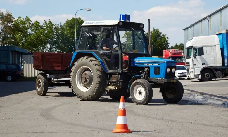 Modrý traktor s přívěsem na asfaltové ploše u průmyslových budov.