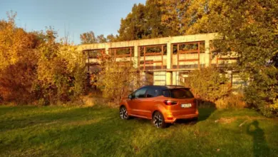 Oranžový Ford EcoSport zaparkovaný na trávníku před opuštěnou budovou.