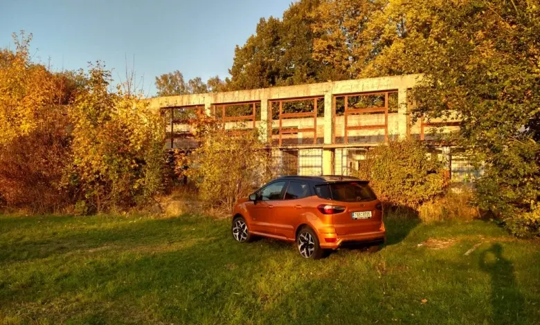 Oranžový Ford EcoSport zaparkovaný na trávníku před opuštěnou budovou.