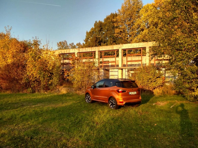 Oranžový Ford EcoSport zaparkovaný na trávníku před opuštěnou budovou.